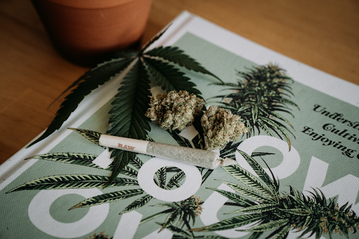 A still-life photograph featuring an unlit joint and a marijuana leaf gracefully arranged on top of a magazine resting on a coffee table. The joint is neatly rolled and awaiting ignition, while the marijuana leaf exhibits its characteristic shape and rich green hues. The image conveys a sense of anticipation and invites contemplation of cannabis culture and its associated relaxation.