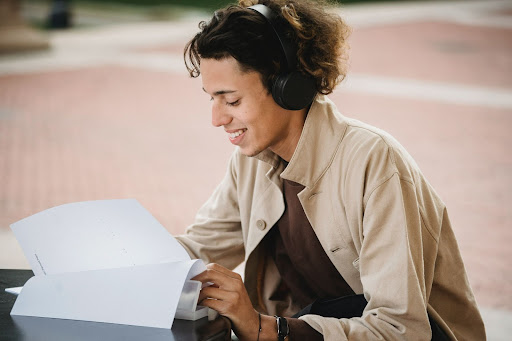 A man wearing headphones, sitting outdoors, with a content smile while reading printed papers.