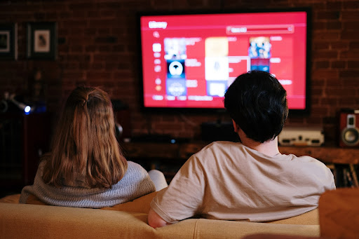 A couple comfortably seated on a couch, attentively watching a large-screen TV.